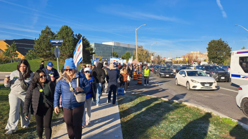 Protestors march down the sidewalk with Humber Polytechnic buildings in the background on a sunny day with cars on the street. 