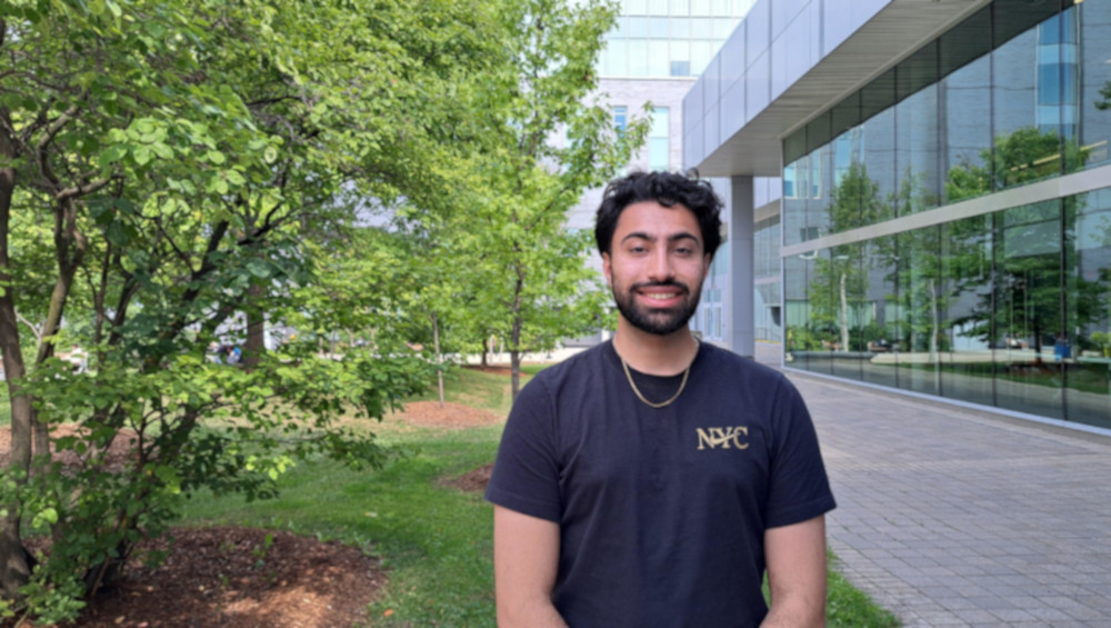 Smiling student standing outside near a modern glass building with trees in the background.