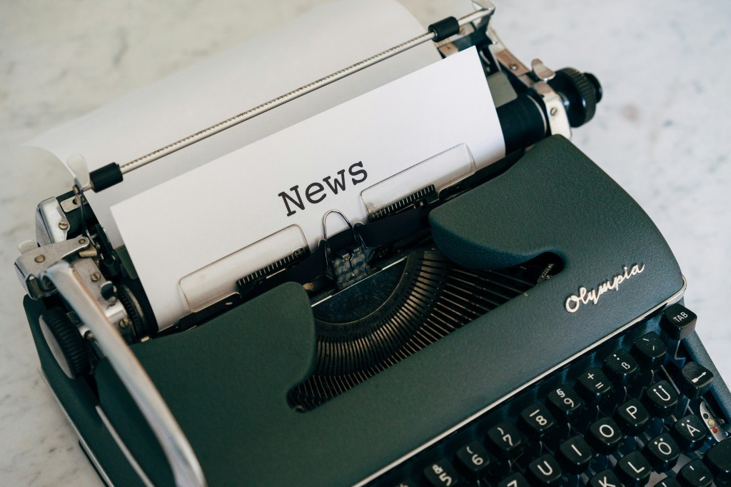 Image of a green Olympia typewriter with a paper in it and the word "News" printed on paper. 